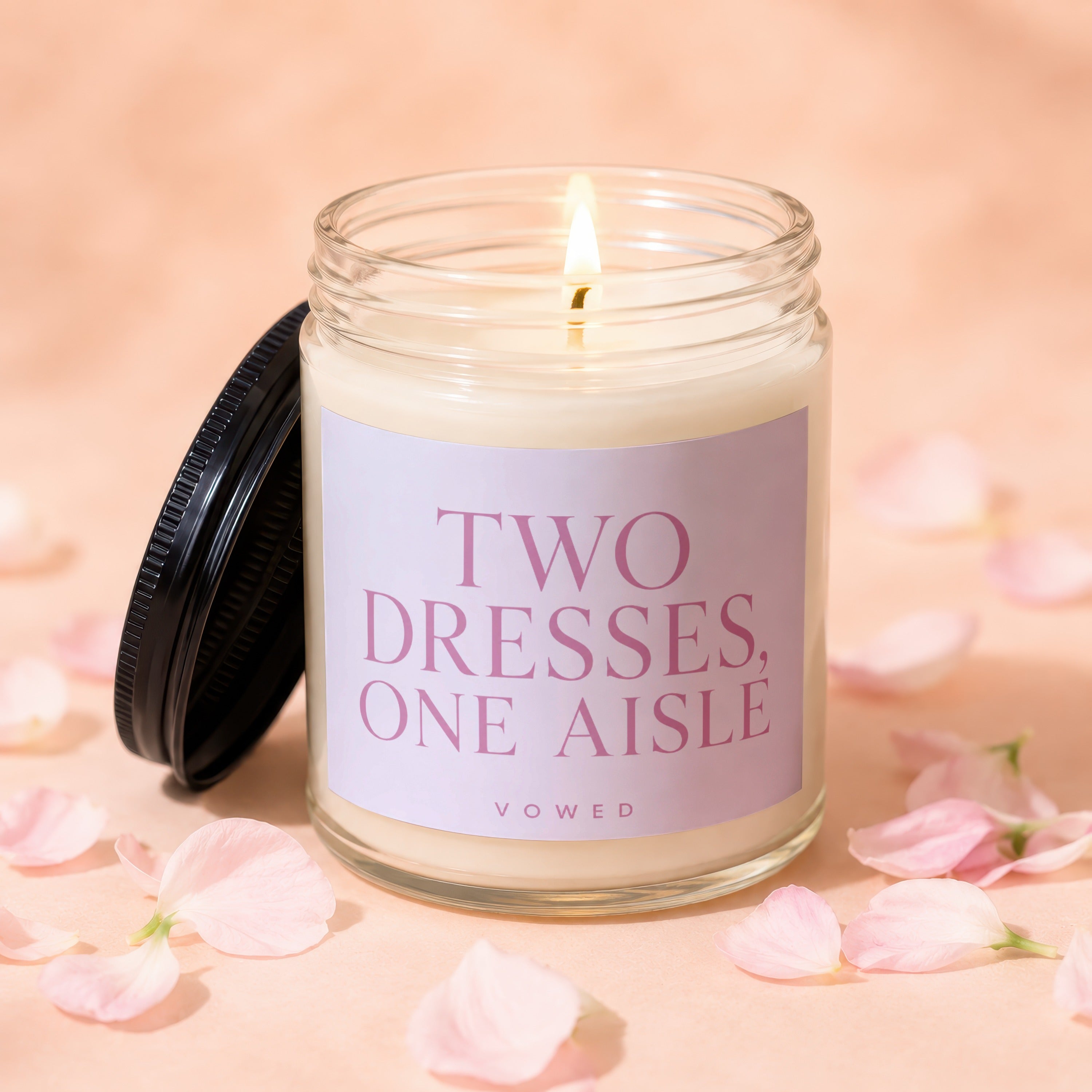 Candle with lit wick, black lid, and pink text reading "TWO DRESSES, ONE AISLE VOWED".