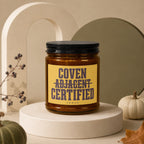 Brown candle in glass jar with black lid. Yellow label reads "Coven Adjacent Certified".