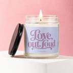 Candle with lit wick and black lid, reads "Love, out loud" and "VOWED".