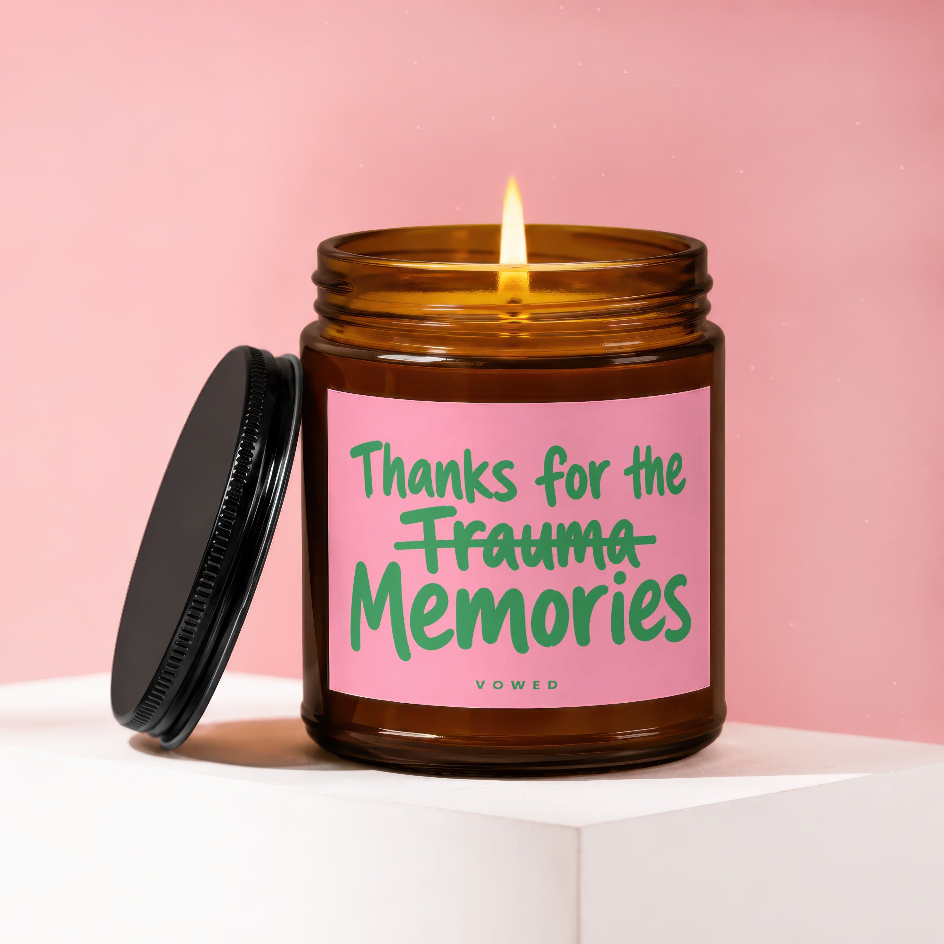 Amber candle with lid off, lit flame, and pink label reading "Thanks for the Trauma Memories".
