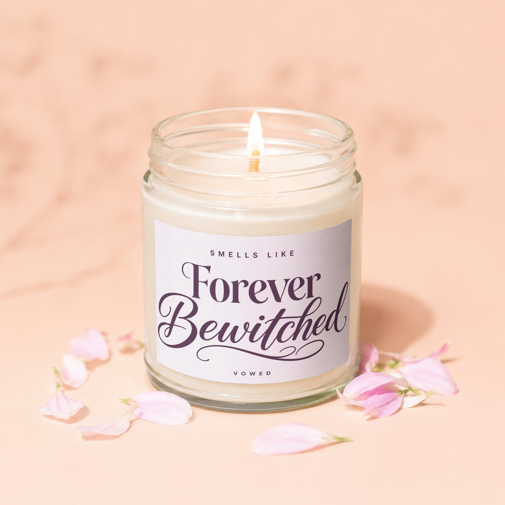 Lit candle in glass jar with "Forever Bewitched" label, surrounded by pink petals.