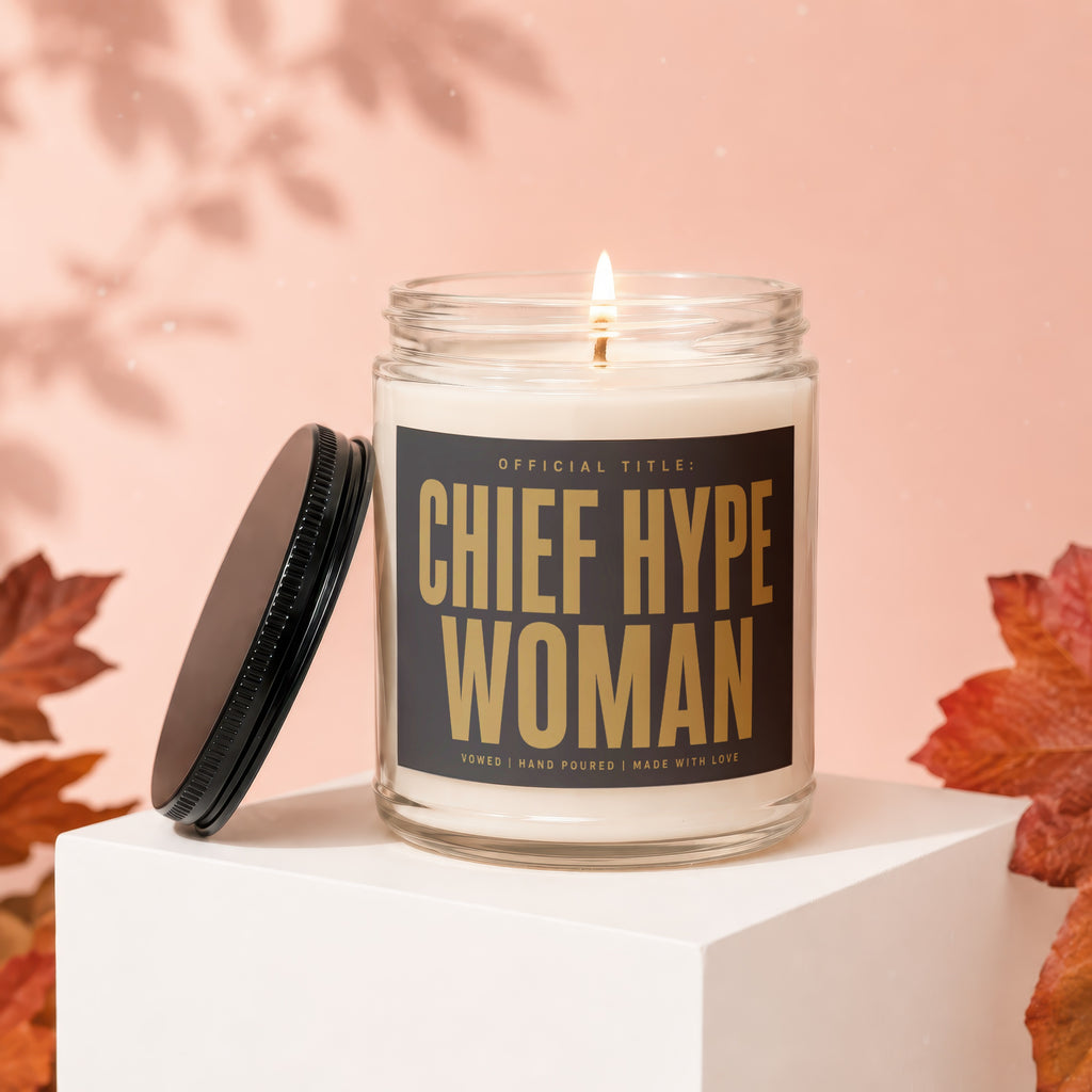 Lit candle with label "Chief Hype Woman" on a white pedestal, surrounded by fall leaves.