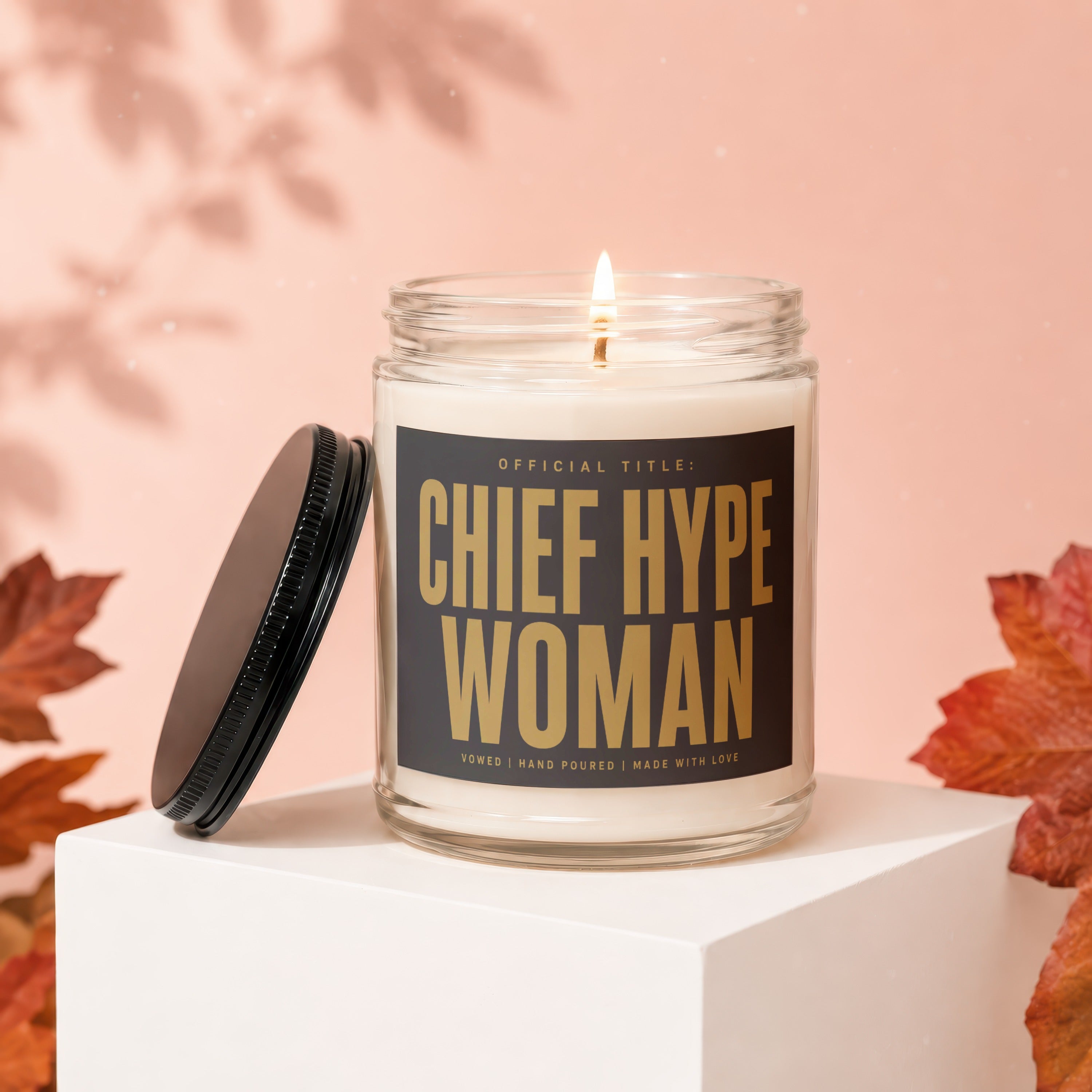 Lit candle with label "Chief Hype Woman" on a white pedestal, surrounded by fall leaves.