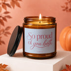 Amber glass candle with lit wick and label saying "So proud of you both"