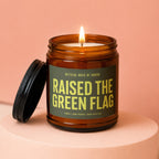 Amber jar candle with lit wick, featuring "RAISED THE GREEN FLAG" text.