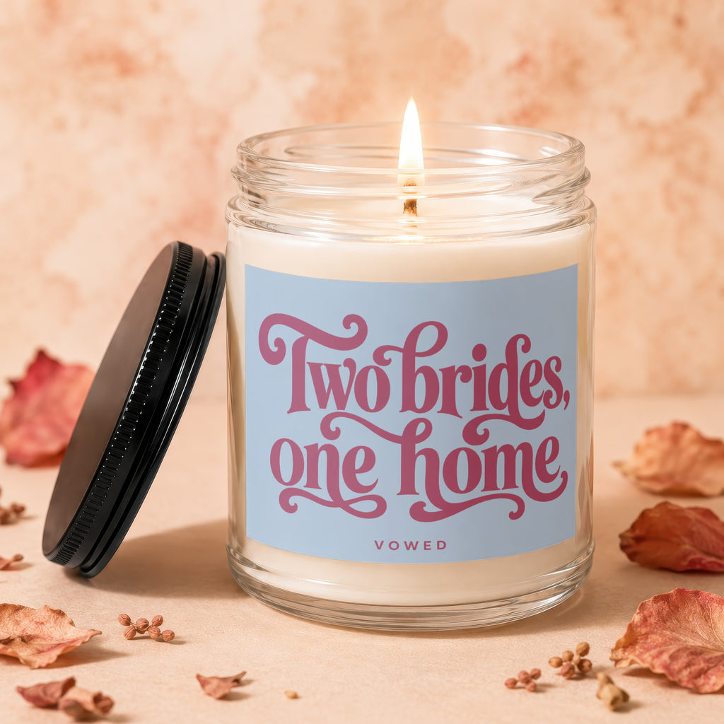 Glass candle with lit wick, label reads "Two brides, one home".