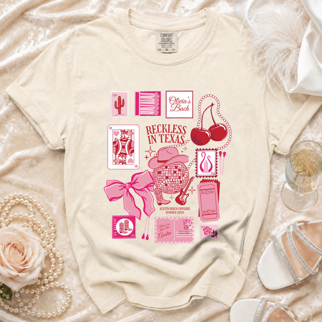 Cream t-shirt with pink graphics: "Olivia's Bach", playing cards, disco ball, cherries, and "Reckless in Texas".