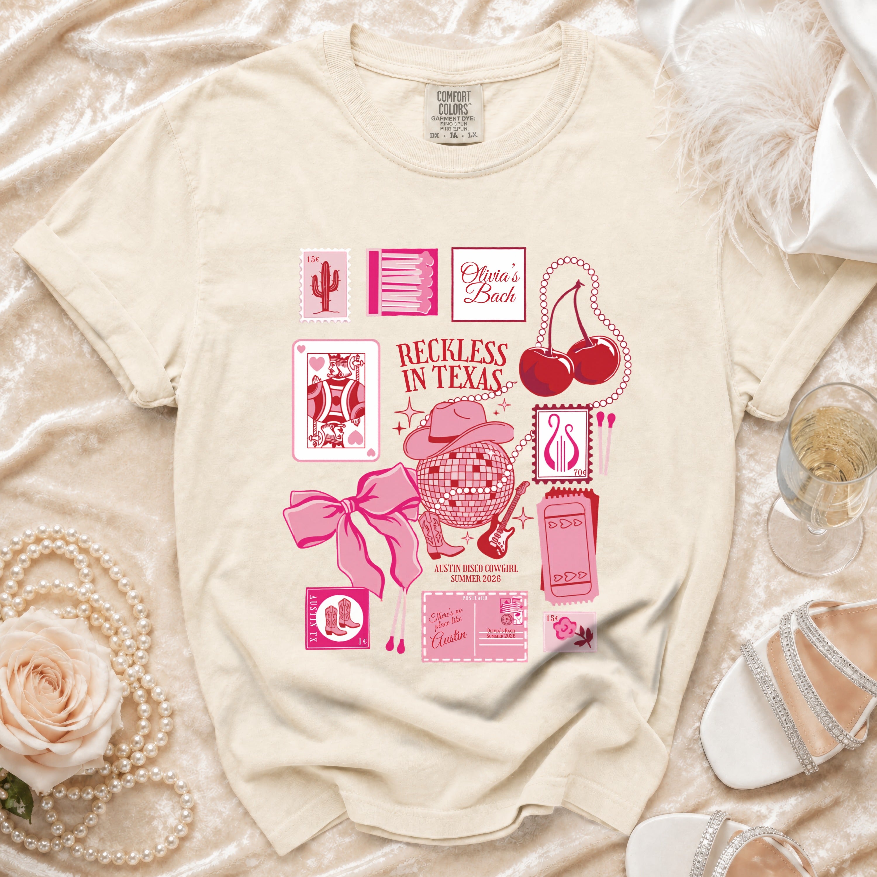 Cream t-shirt with pink graphics: "Olivia's Bach", playing cards, disco ball, cherries, and "Reckless in Texas".