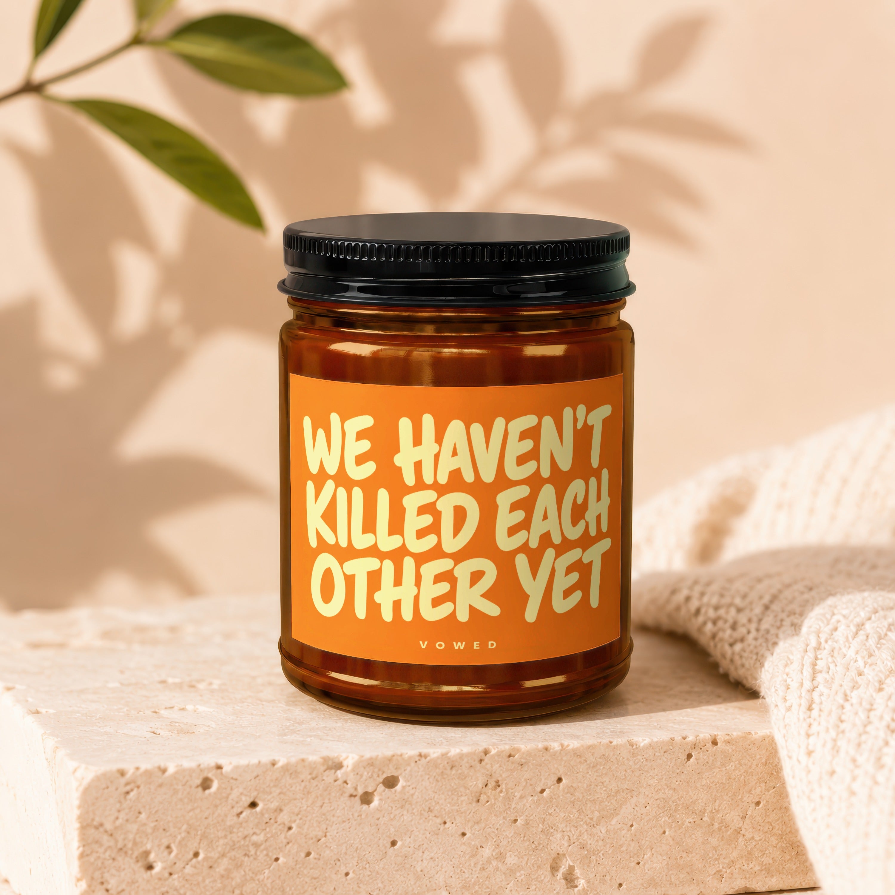 Amber jar candle with black lid featuring text "WE HAVEN'T KILLED EACH OTHER YET."