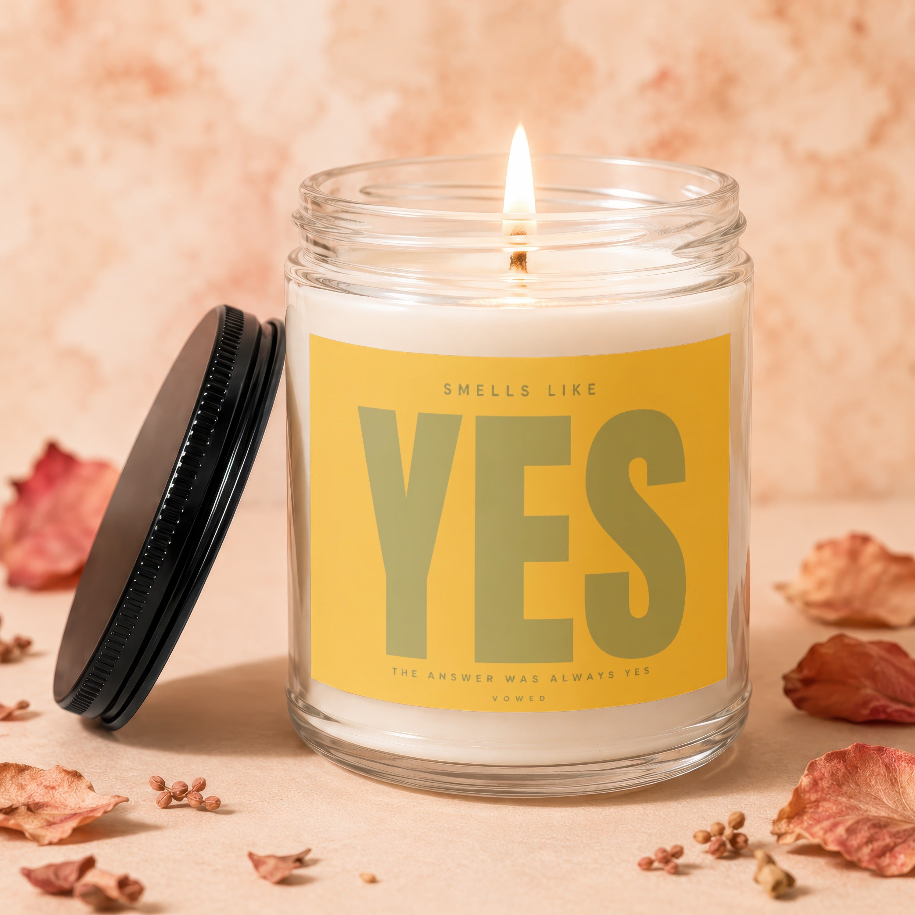 Glass jar candle with lit wick, yellow label reads "SMELLS LIKE YES THE ANSWER WAS ALWAYS YES VOWED".