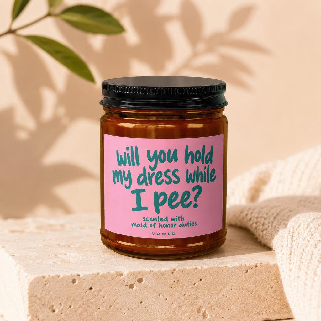 Candle with pink label reading "will you hold my dress while I pee?"