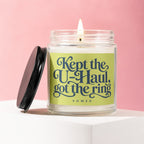 Lit candle in a glass jar with a black lid, displaying "Kept the U-Haul, got the ring" text.