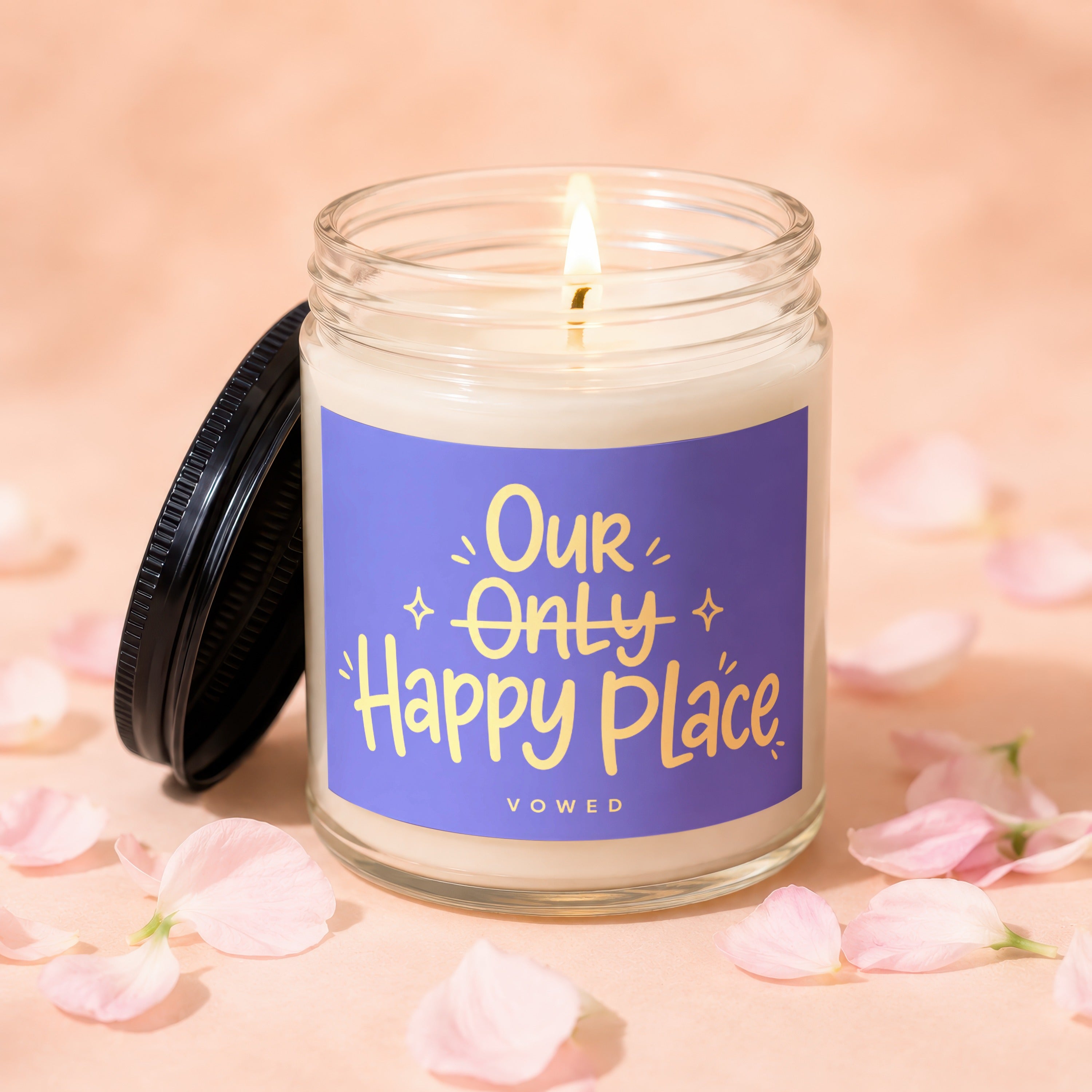 Lit candle in jar with label reading "Our Only Happy Place VOWED" surrounded by petals.