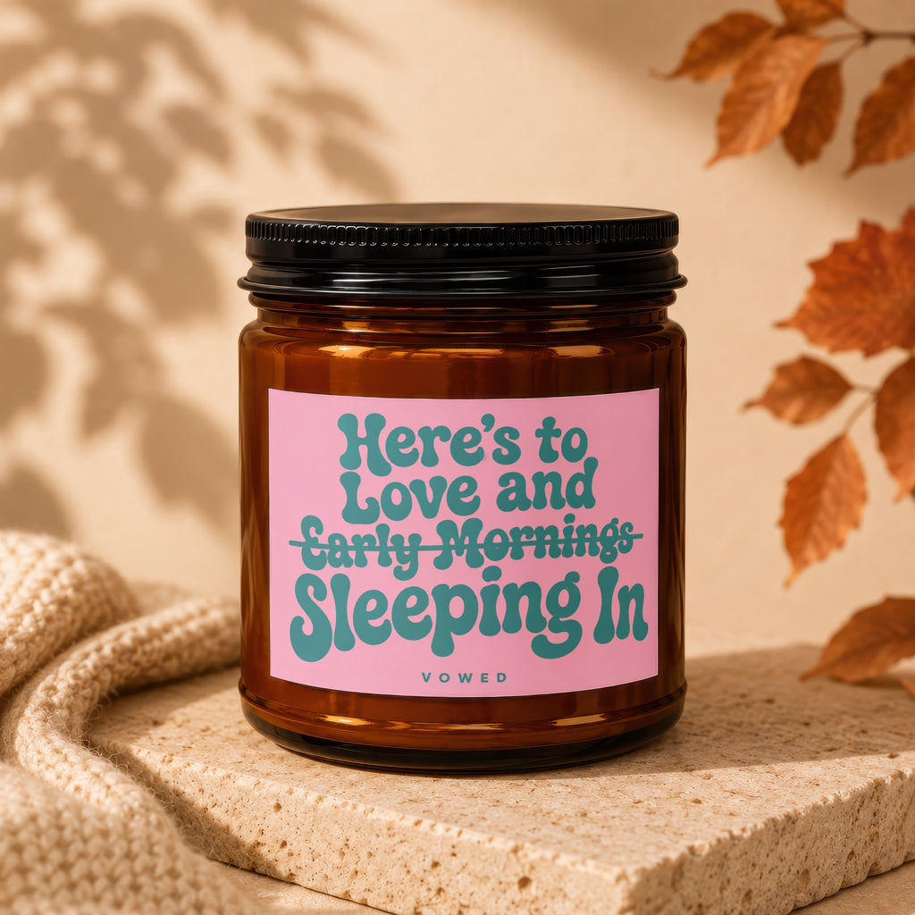Amber glass candle with pink label reading "Here's to Love and Early Mornings Sleeping In".