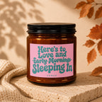 Amber glass candle with pink label reading "Here's to Love and Early Mornings Sleeping In".