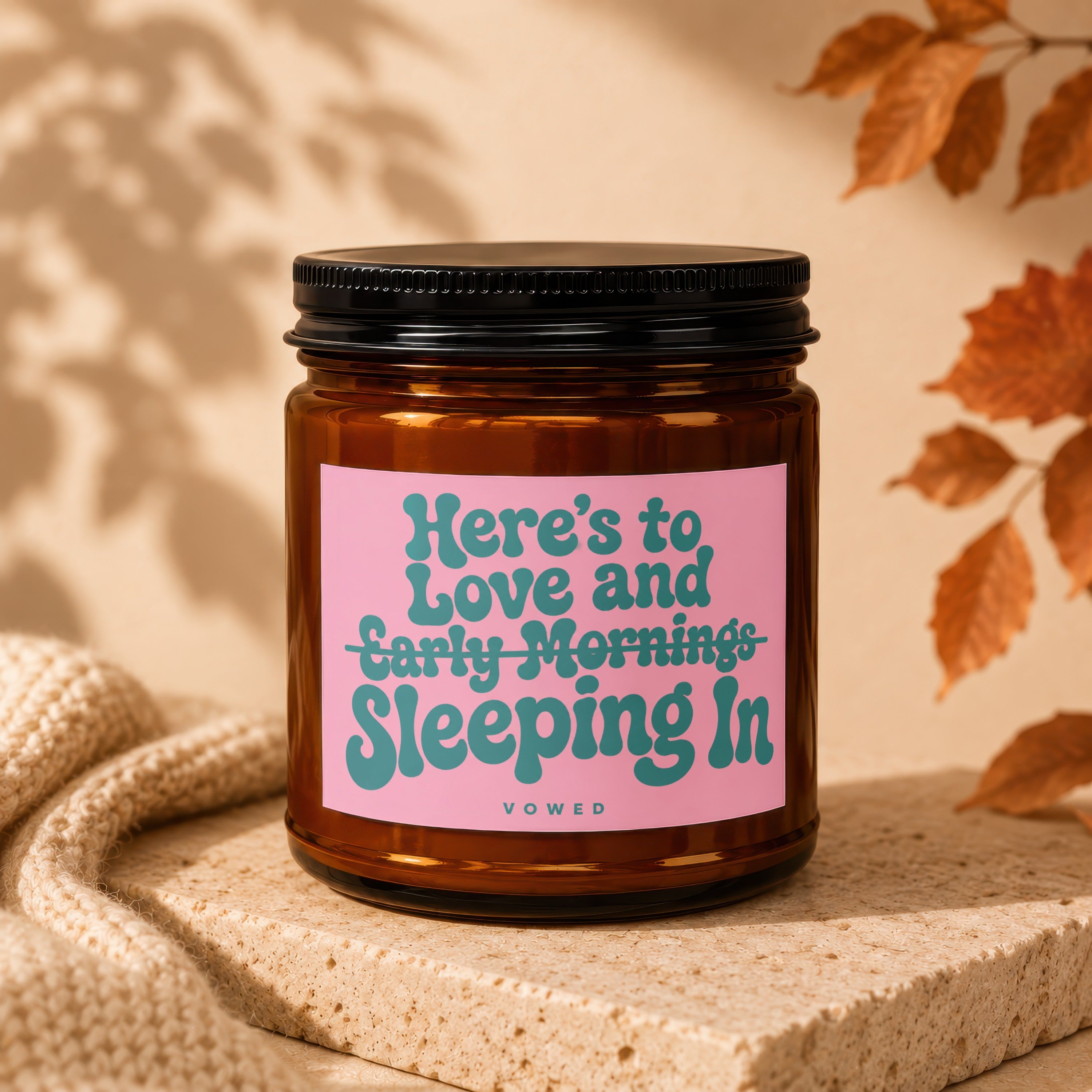 Amber glass candle with pink label reading "Here's to Love and Early Mornings Sleeping In".