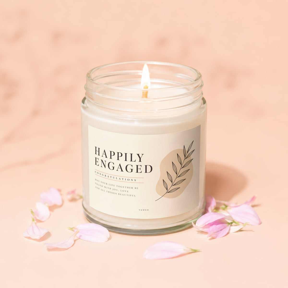 Glass candle with lit wick, "Happily Engaged" label, and scattered pink flower petals.