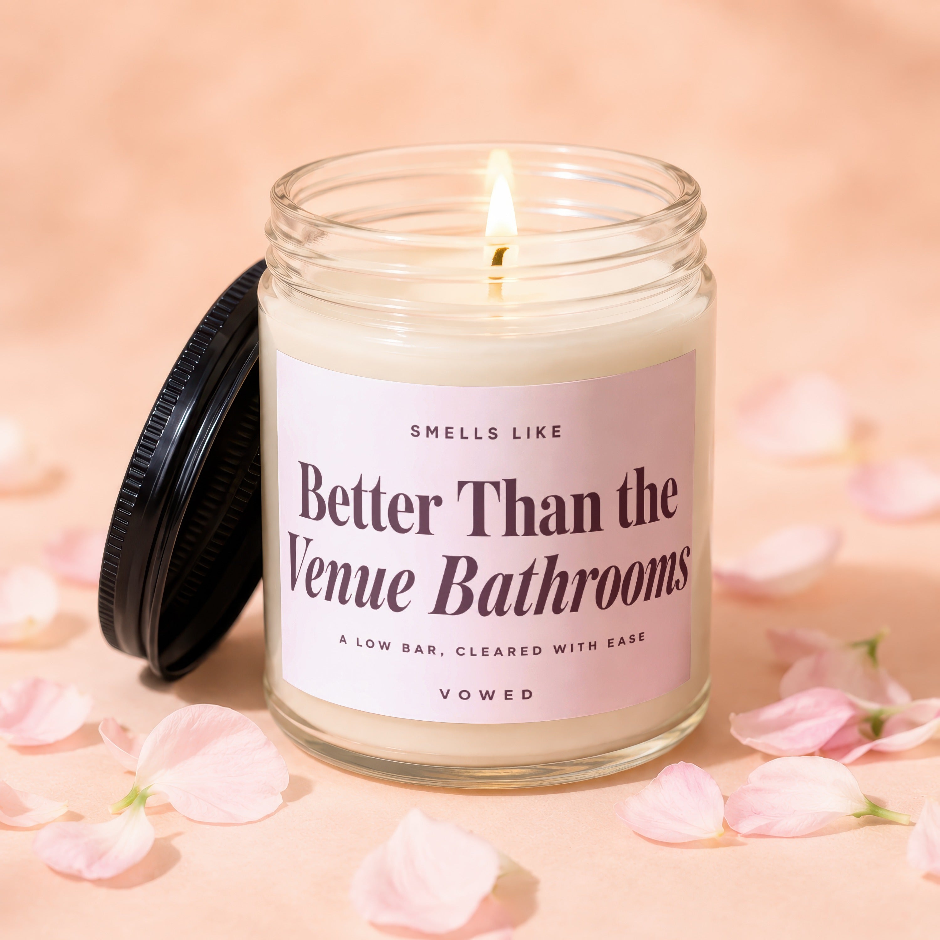Vowed candle, "Better Than the Venue Bathrooms" scent, on a pink background with petals.