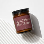 Glass candle jar with maroon label reading "Second Time's the Charm" and "VOWED".