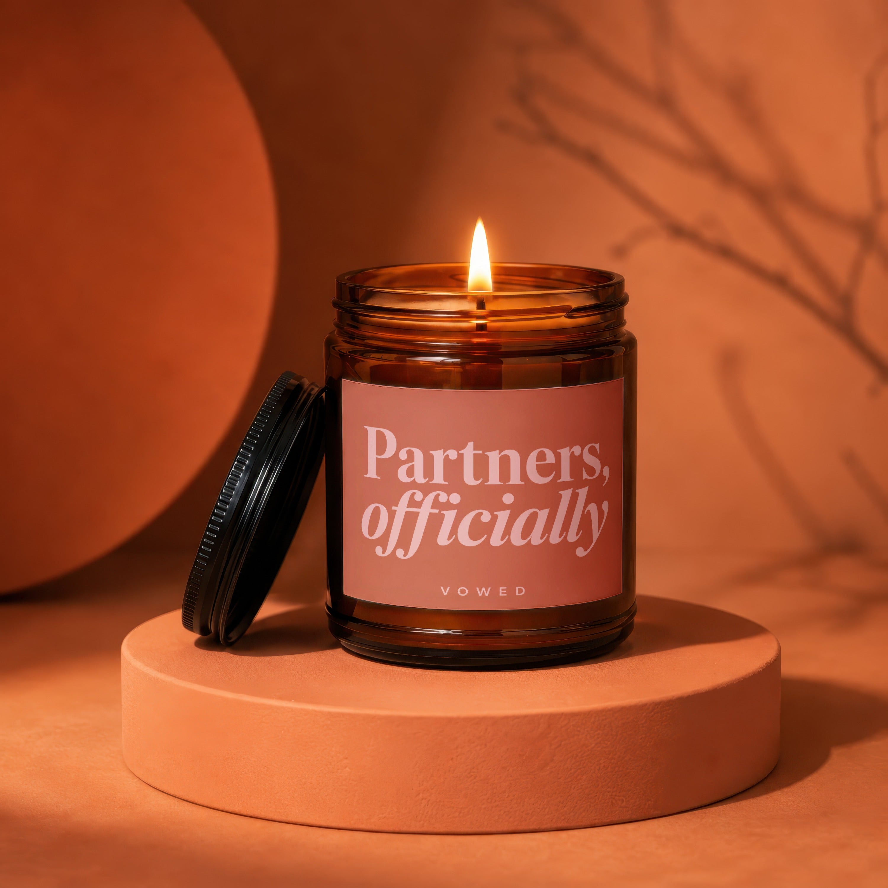 Lit amber glass candle with "Partners, officially VOWED" label on coral pedestal.