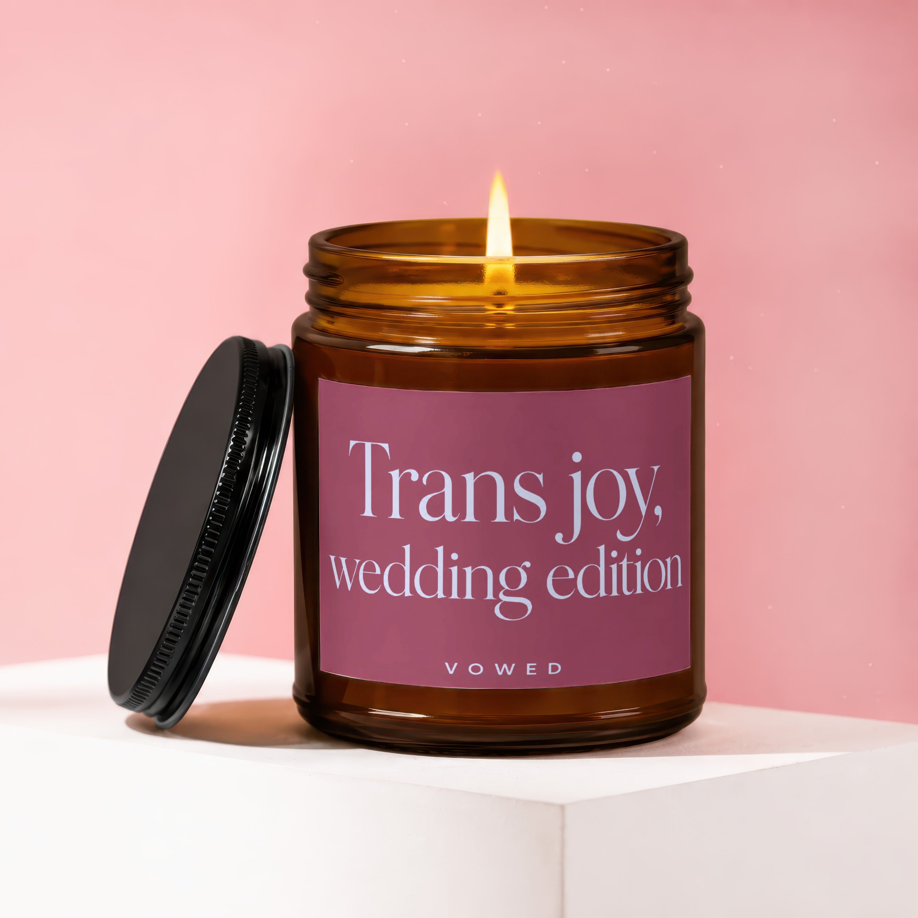 A lit amber glass candle with a pink label reading "Trans joy, wedding edition".