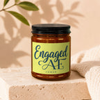 Amber jar candle with black lid and bright yellow label reading "Engaged AF VOWED".