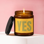 Amber candle with lit wick, yellow label reads "SMELLS LIKE YES THE ANSWER WAS ALWAYS YES VOWED".