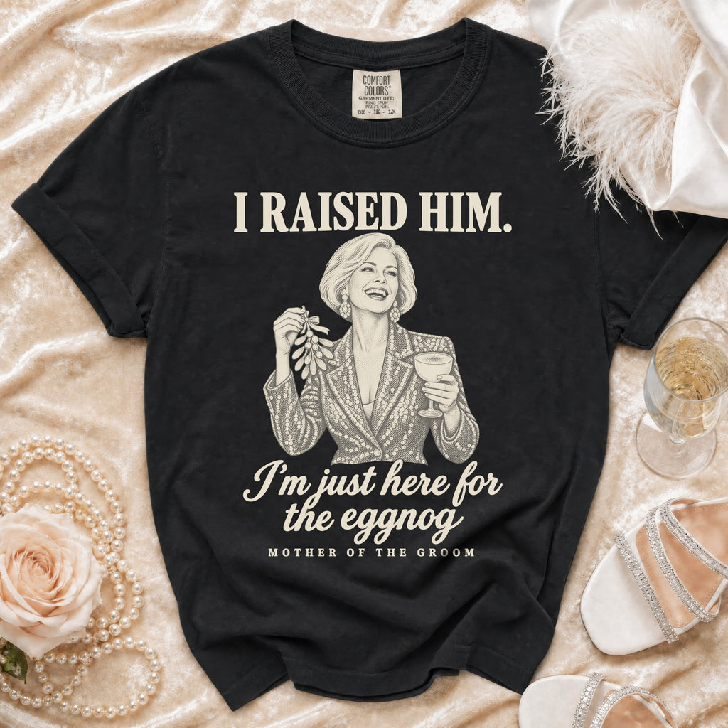Black t-shirt featuring a graphic illustration of a woman holding mistletoe and eggnog with text reading I RAISED HIM. I'm just here for the eggnog. MOTHER OF THE GROOM.