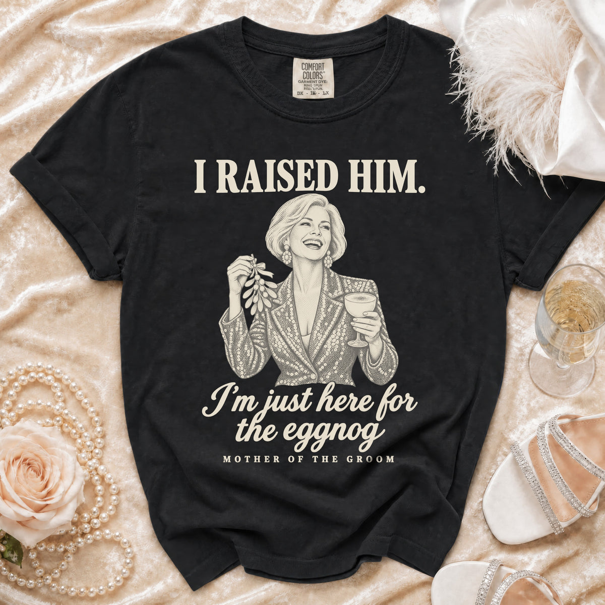 Black t-shirt featuring a graphic illustration of a woman holding mistletoe and eggnog with text reading I RAISED HIM. I'm just here for the eggnog. MOTHER OF THE GROOM.