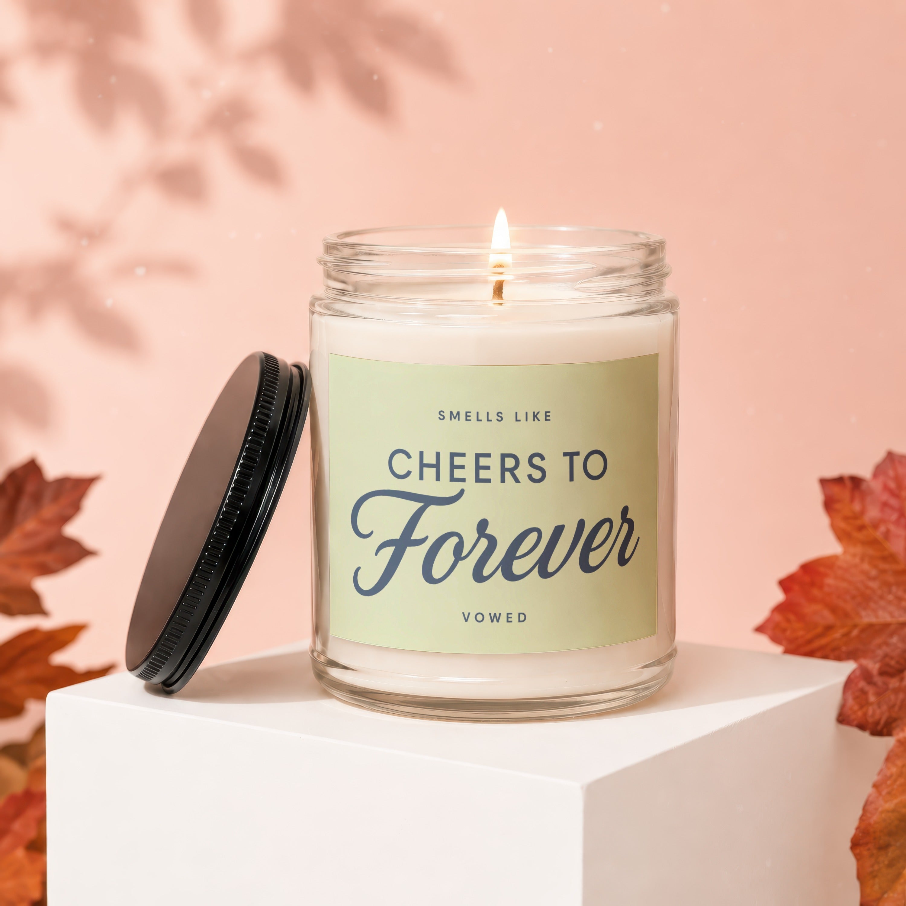 Glass candle with label that reads "SMELLS LIKE Cheers To Forever" on a white pedestal.