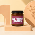 Amber jar candle with a maroon label reading "Our Forever Begins".