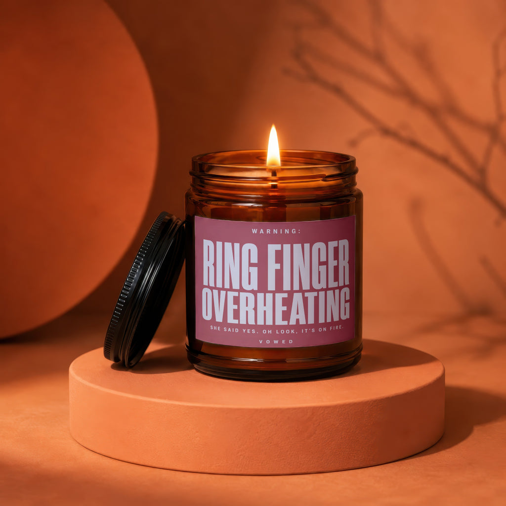 Candle with label "Ring Finger Overheating" in amber glass jar with lit wick.