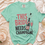 Aqua t-shirt with text "THIS BRIDE NEEDS CHAMPAGNE", bottle, and glass.