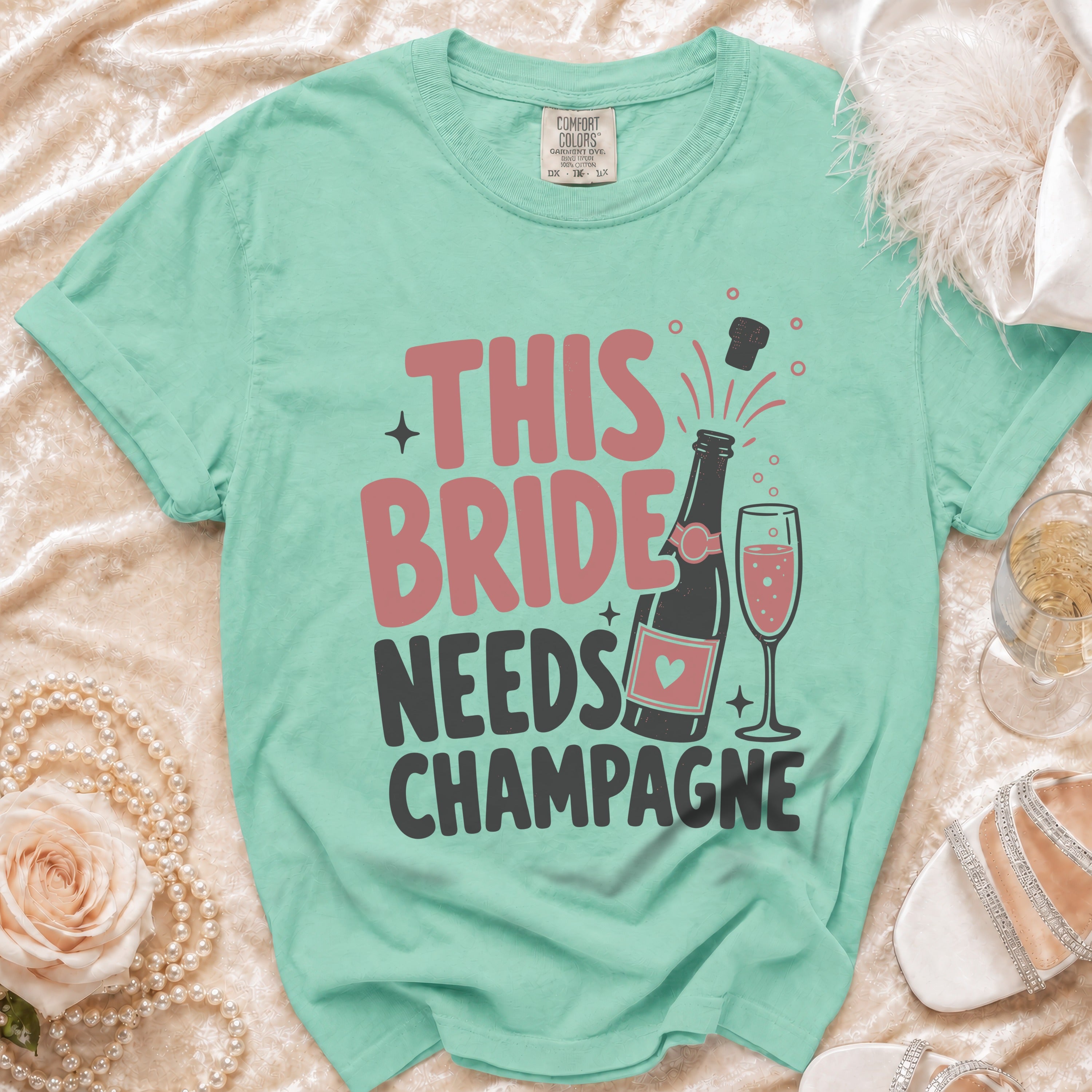 Aqua t-shirt with text "THIS BRIDE NEEDS CHAMPAGNE", bottle, and glass.