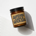 Amber glass candle with black lid reads "Smells like Someone Else's Open Bar".