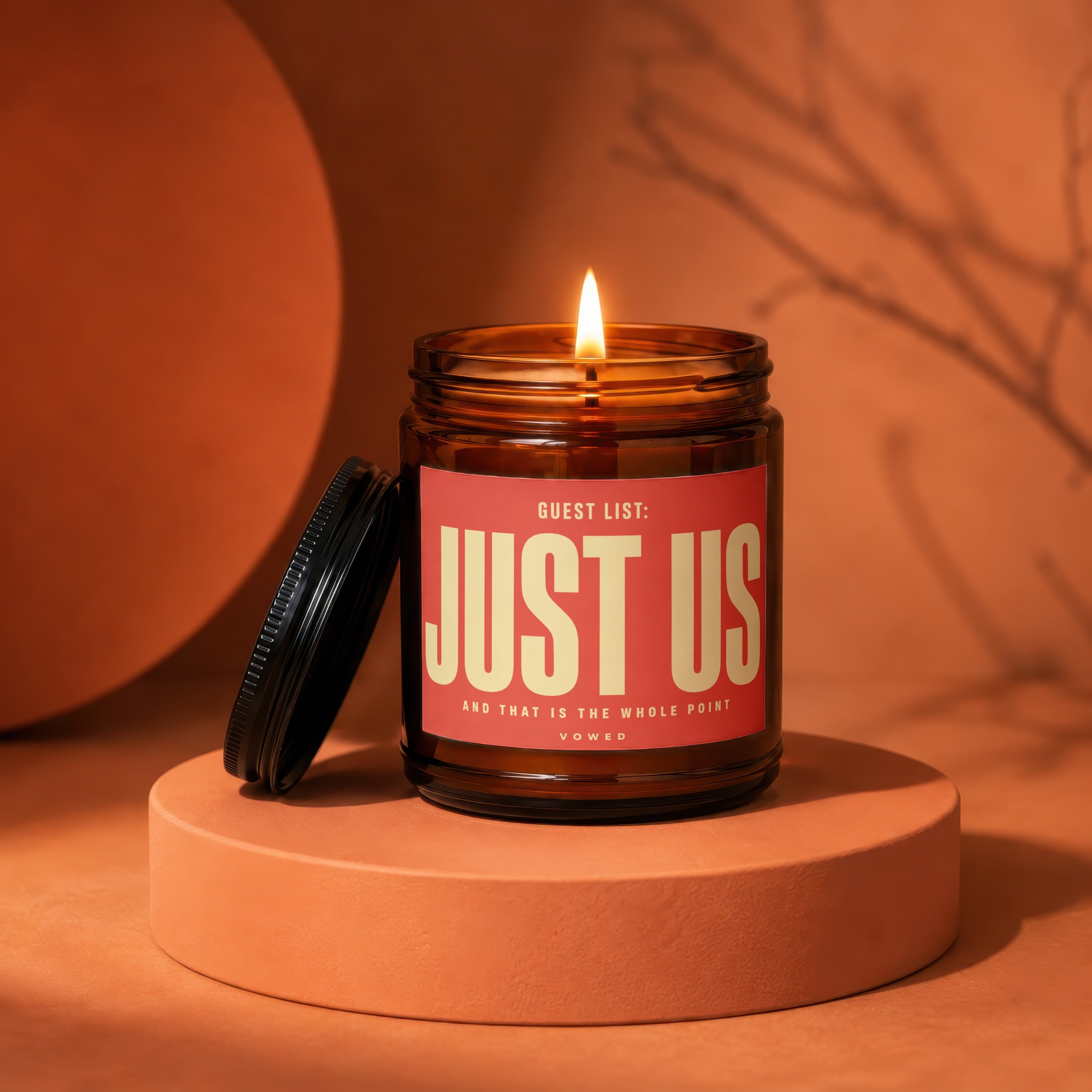 Amber candle with lit wick, label reads "GUEST LIST: JUST US".