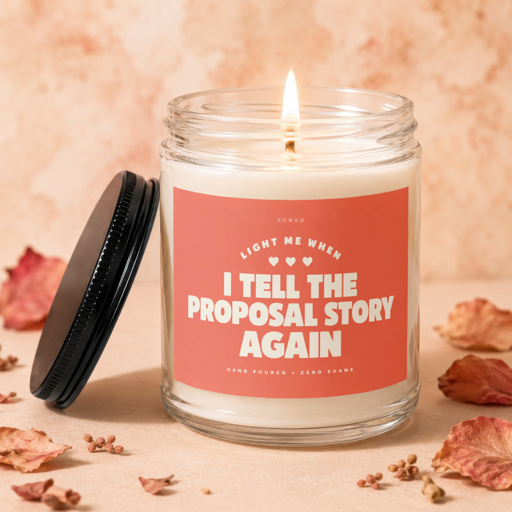 Glass candle with lit wick and coral label that reads "Light me when I tell the proposal story again."
