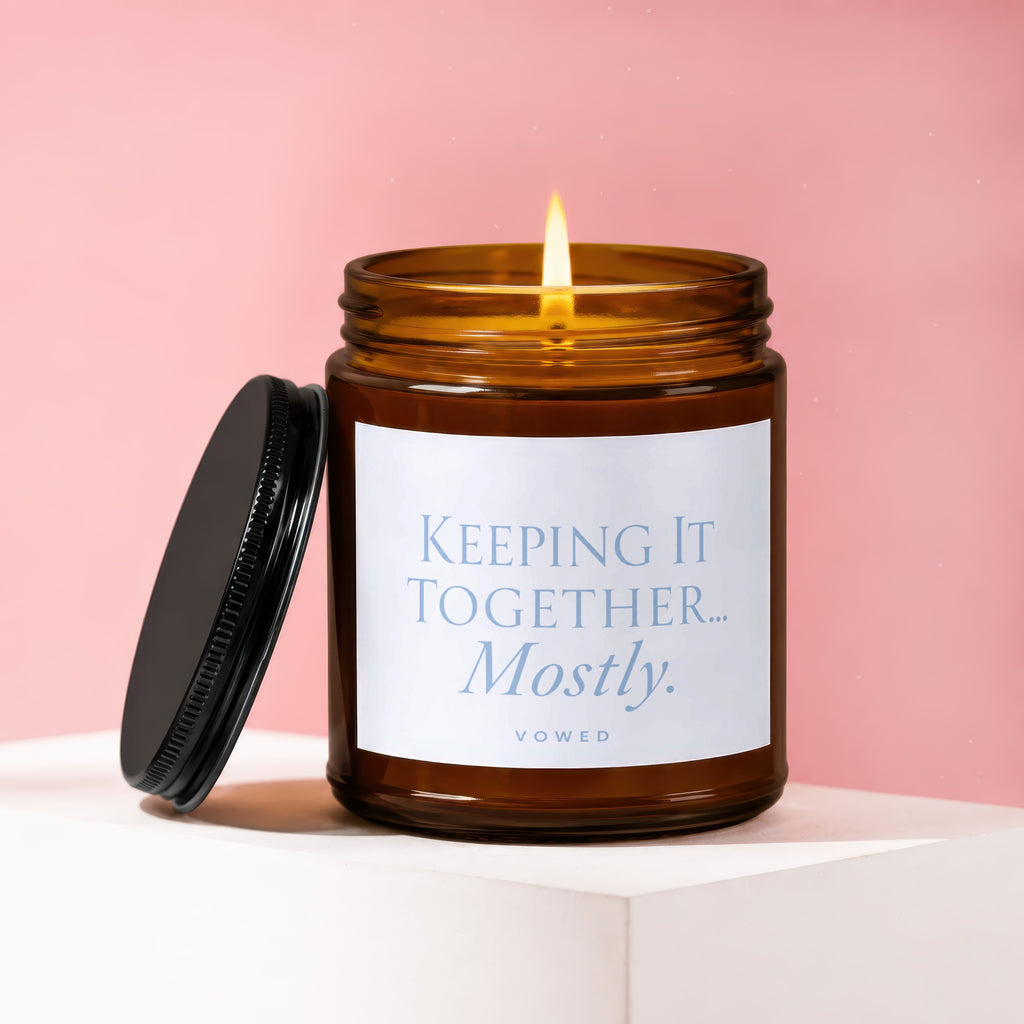 Amber glass candle burning with a white label saying "Keeping It Together... Mostly. VOWED."