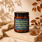 A brown glass candle with a dark lid printed with "Will you be my maid of honor? Act like this is a surprise."