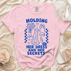 Pink t-shirt with blue graphic of dress, champagne, and text "HOLDING HER DRESS AND HER SECRETS".