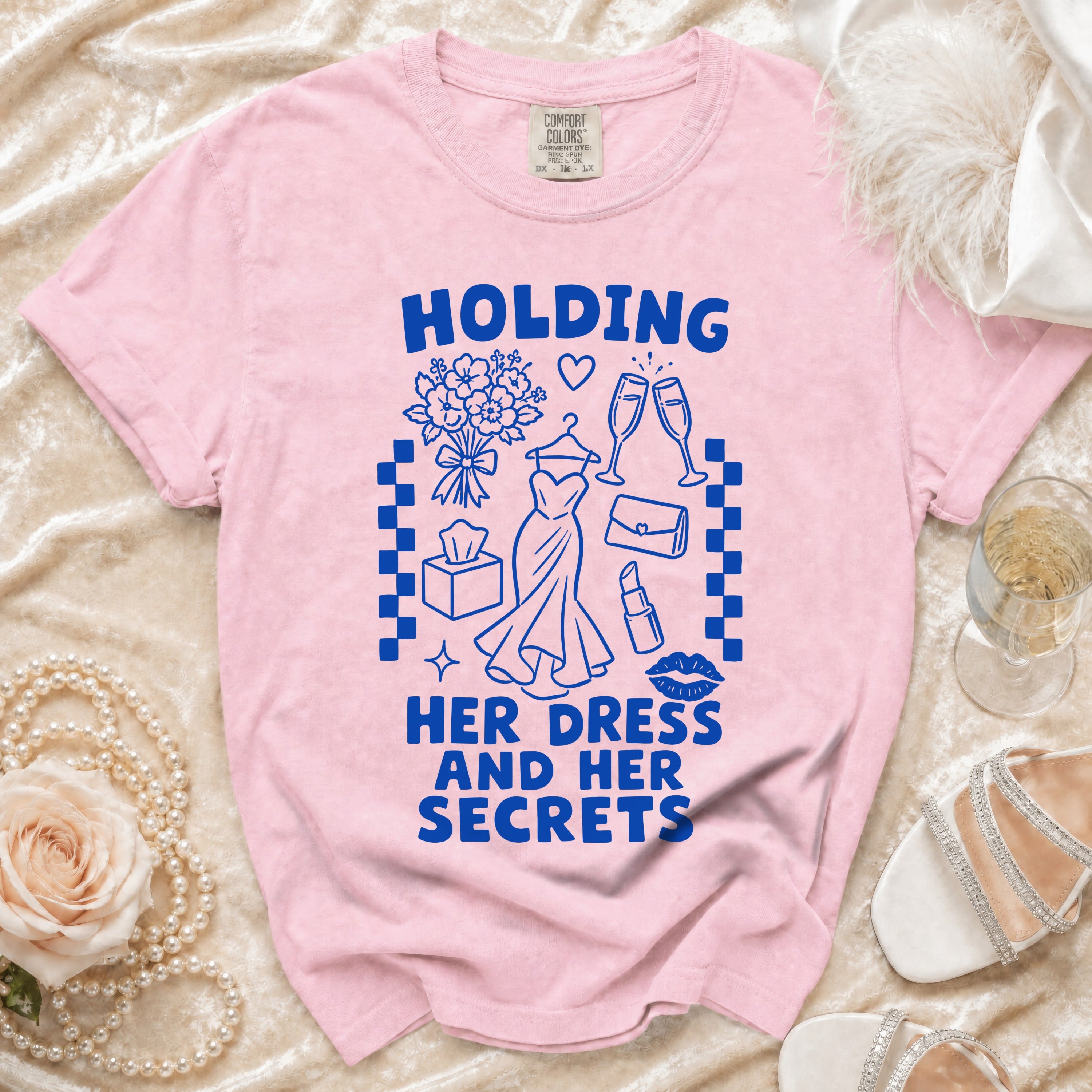 Pink t-shirt with blue graphic of dress, champagne, and text "HOLDING HER DRESS AND HER SECRETS".