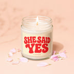 Glass jar candle with lit wick, label reads "SHE SAID YES VOWED", surrounded by pink petals.