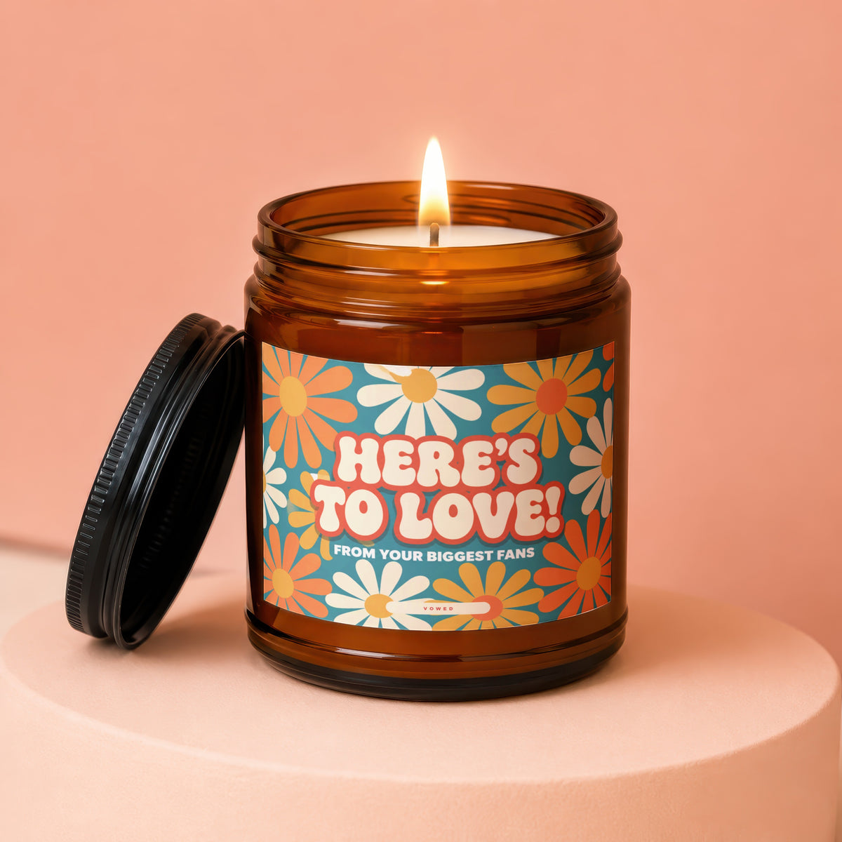 Amber jar candle with lit flame and floral label reading "HERE'S TO LOVE! FROM YOUR BIGGEST FANS".