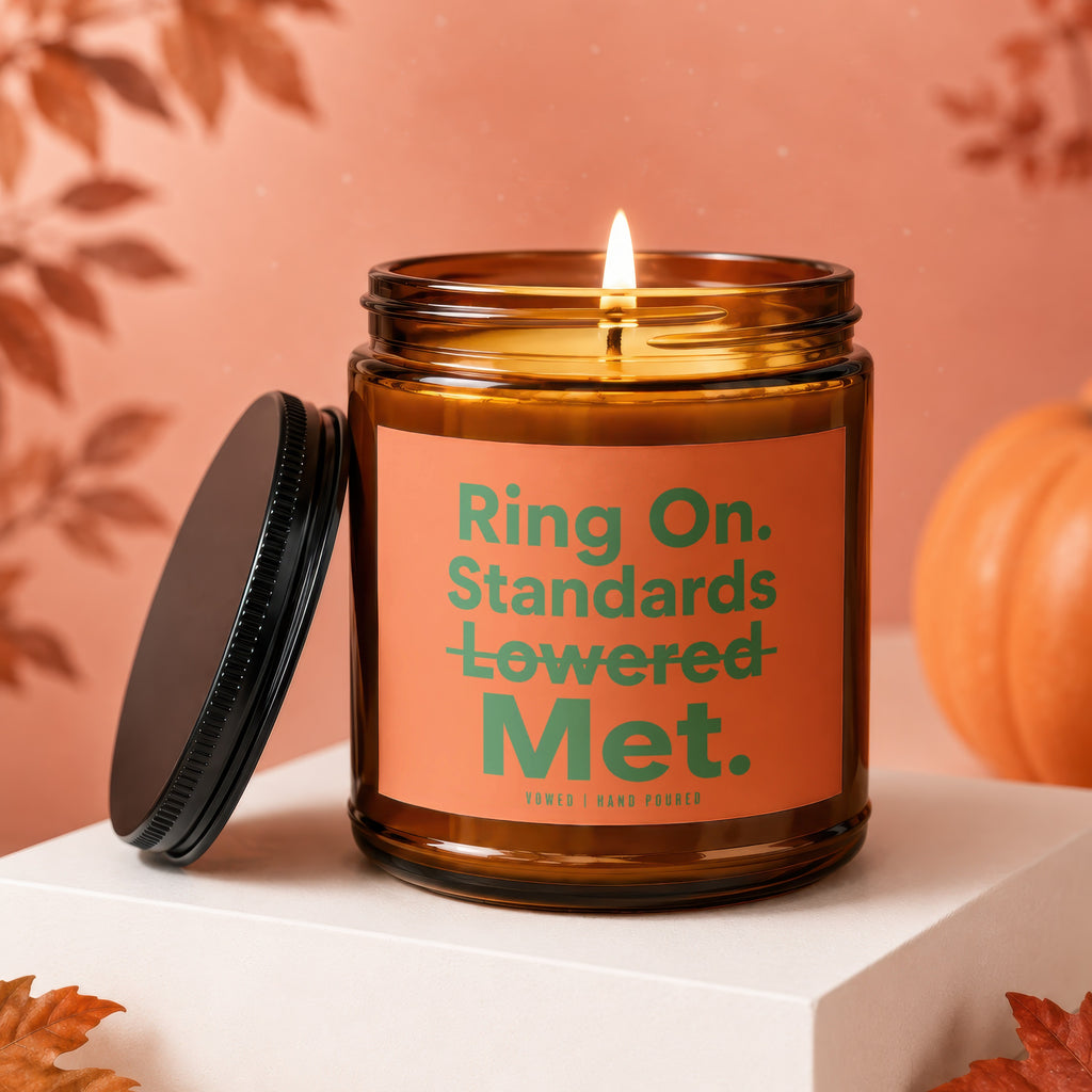 Amber jar candle with lit flame and black lid, featuring "Ring On. Standards Lowered Met." text.