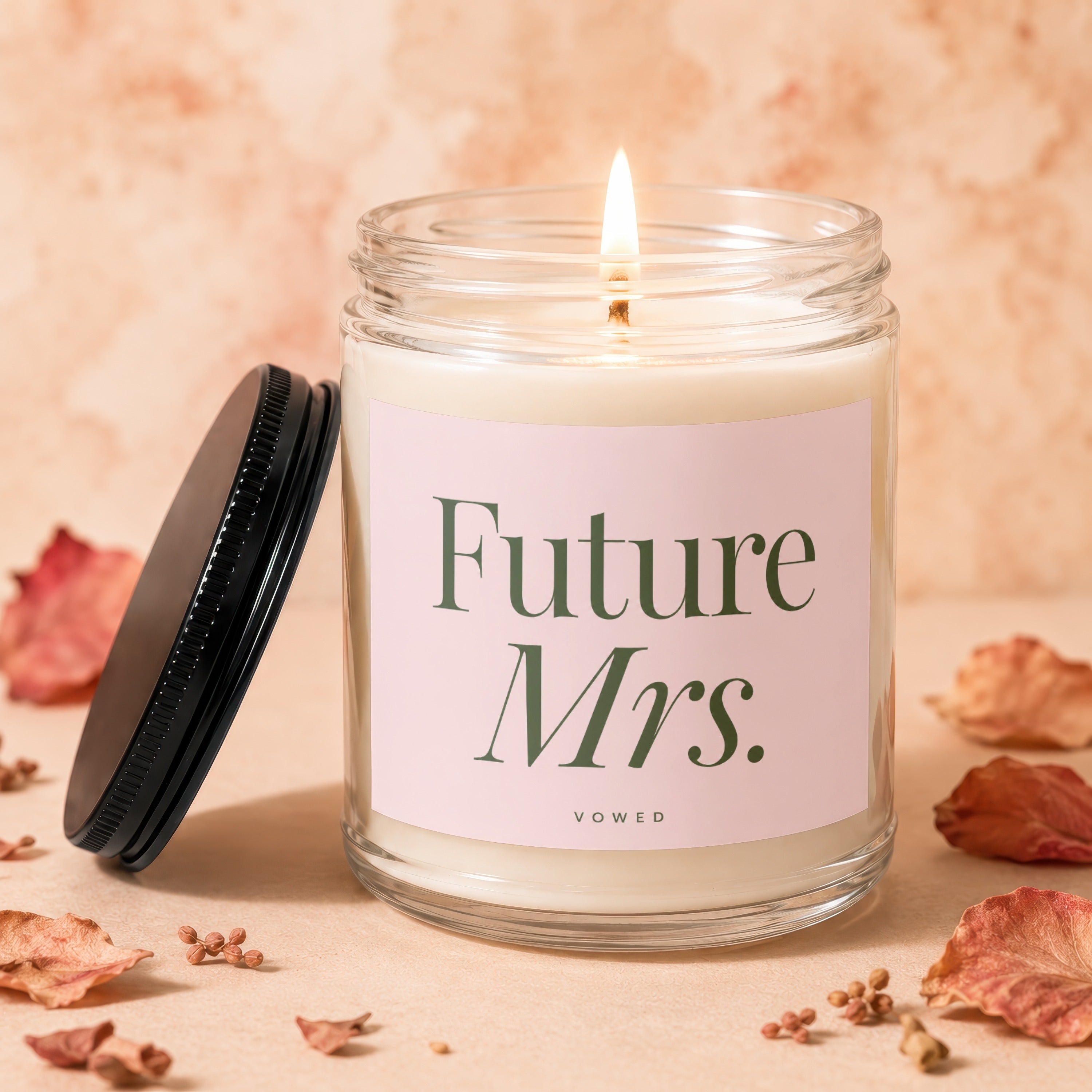Lit candle in clear jar with "Future Mrs." text on pink label, black lid beside it.