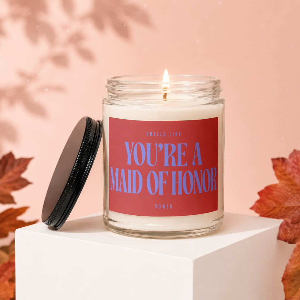 Lit candle in jar with red label that reads "SMELLS LIKE YOU'RE A MAID OF HONOR".