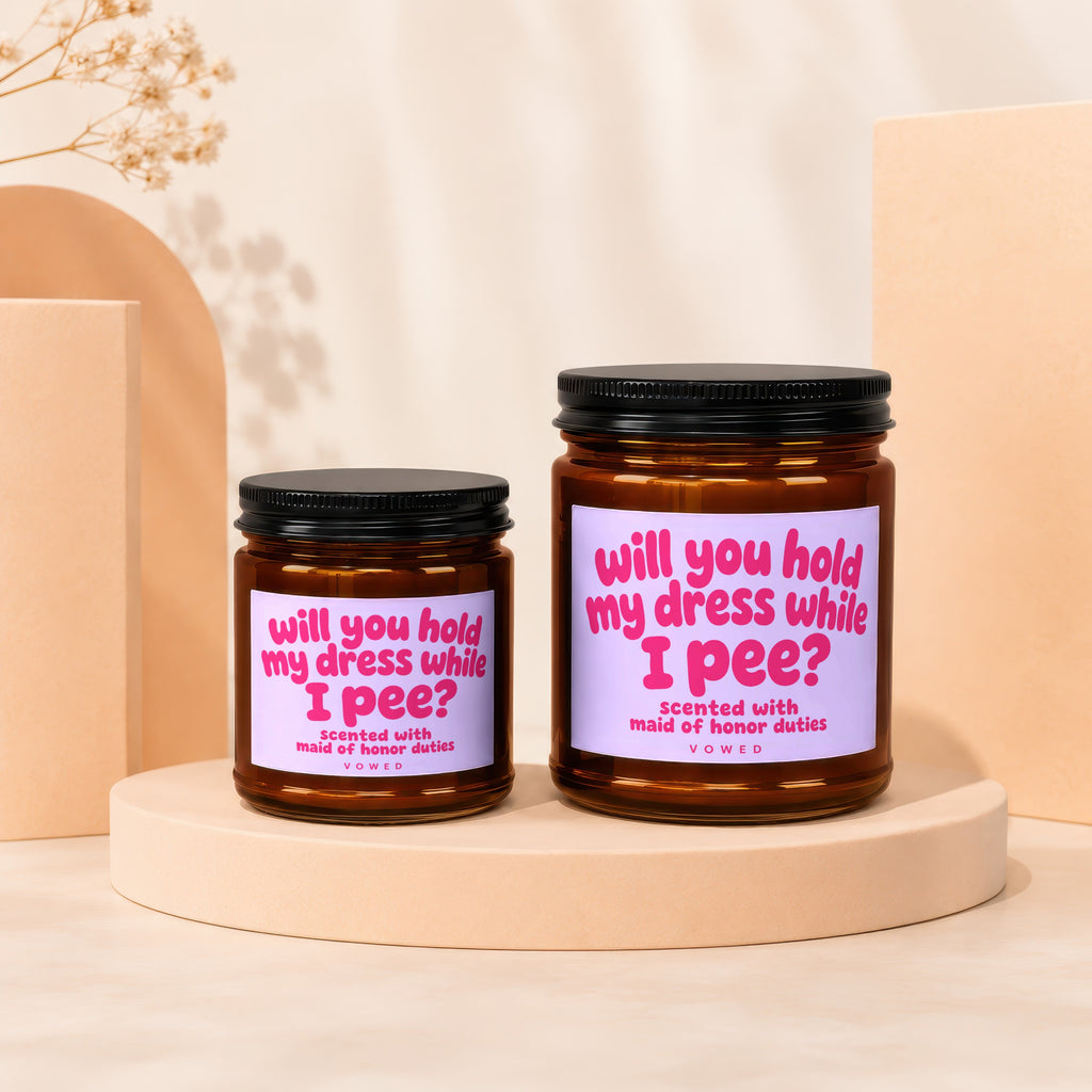 Two amber glass candles with lavender labels reading "Will you hold my dress while I pee?"