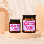 Two amber glass candles with lavender labels reading "Will you hold my dress while I pee?"