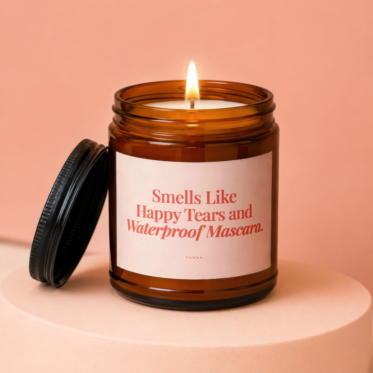 Amber glass candle with lit flame, "Smells Like Happy Tears and Waterproof Mascara." text.