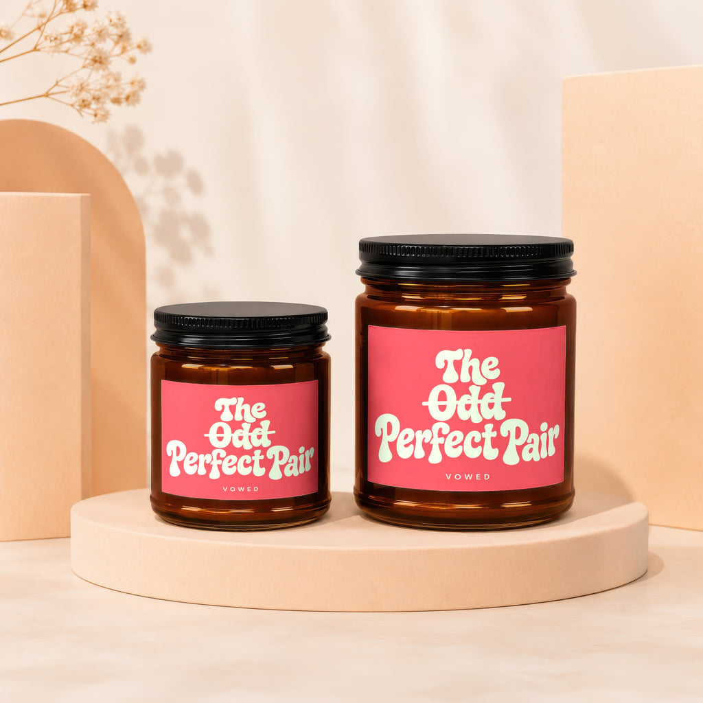 Two amber glass candles with pink labels that say "The Odd Perfect Pair Vowed".